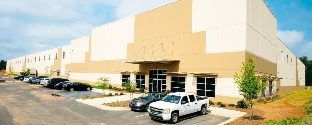 DSCF6062_small A modern beige and white warehouse with parked cars in front, featuring a large glass entrance and landscaping.