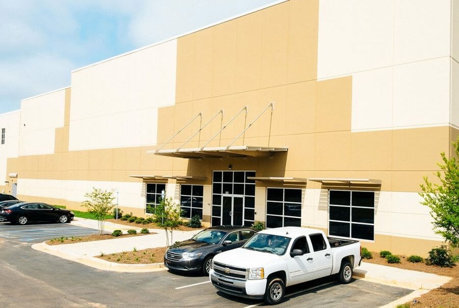 A modern beige and white warehouse with parked cars in front, featuring a large glass entrance and landscaping.