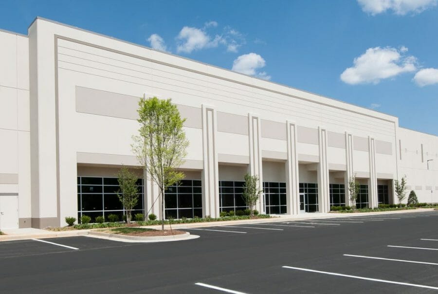 Modern warehouse exterior with large windows, an empty parking lot, cloudy sky, and minimal landscaping.