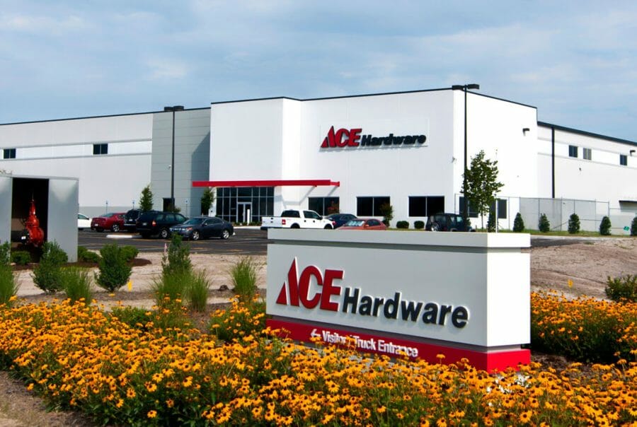 Ace Hardware distribution center with sign and yellow flowers in front, cars parked, and modern building in background.