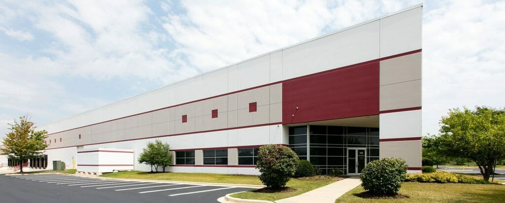 475-copy_small Modern industrial building exterior with red accents, glass entrance, landscaped shrubs, and an empty parking lot.