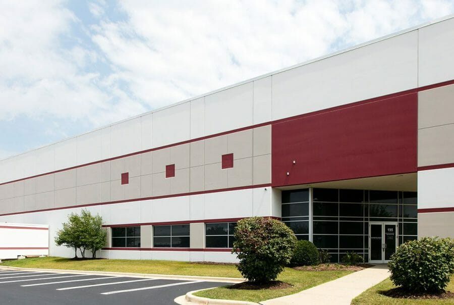 Modern industrial building exterior with red accents, glass entrance, landscaped shrubs, and an empty parking lot.