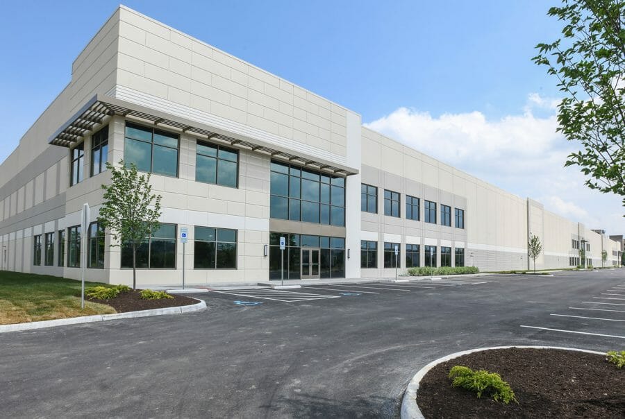 Modern industrial warehouse exterior with large windows, expansive parking lot, clear blue sky, and minimal landscaping.