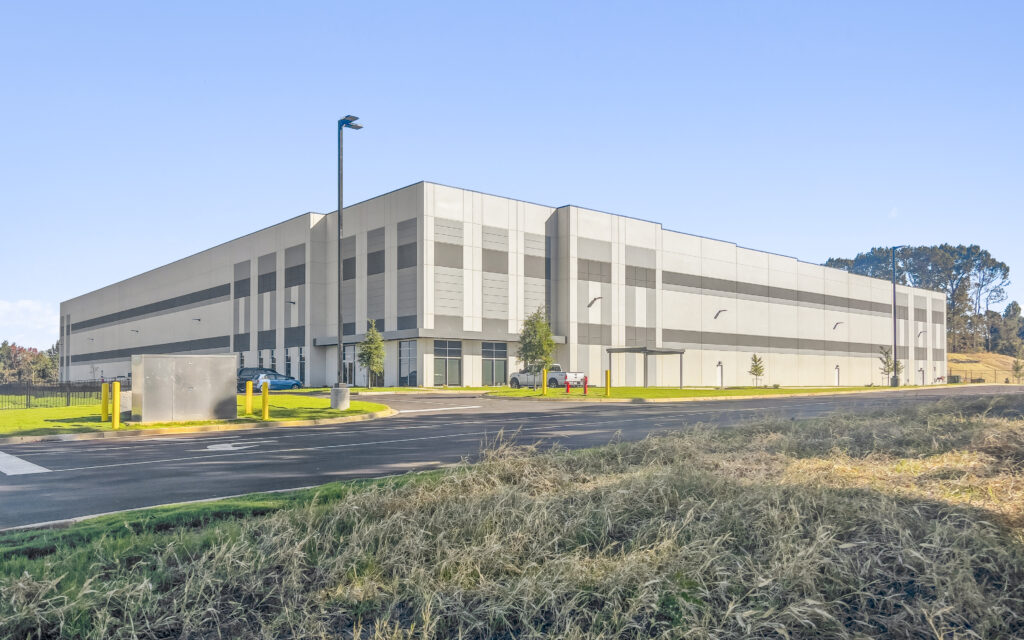 Modern industrial warehouse building with a gray facade, surrounded by grass and a paved road under clear blue sky.