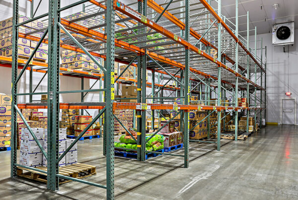 Warehouse interior showing stocked shelves with organized boxes and pallets on a clean concrete floor, under bright lighting.