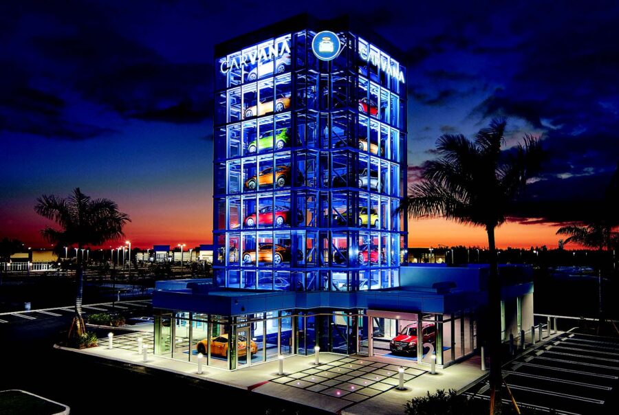 Car vending machine tower lit up at dusk, showcasing colorful cars in glass display, surrounded by palm trees against sunset sky.